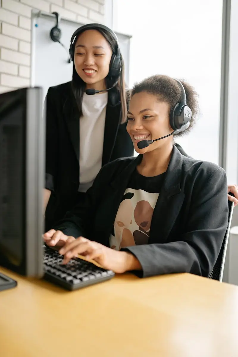 Laughing call center agents wearing headsets collaborate on a computer task in a modern office.